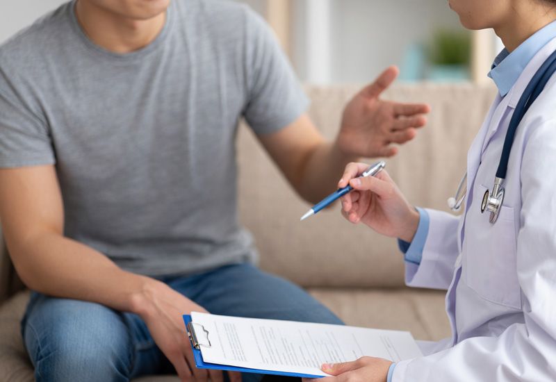 Мужчина в серой футболке и джинсах на домашнем приеме врача нарколога с планшетом в руках